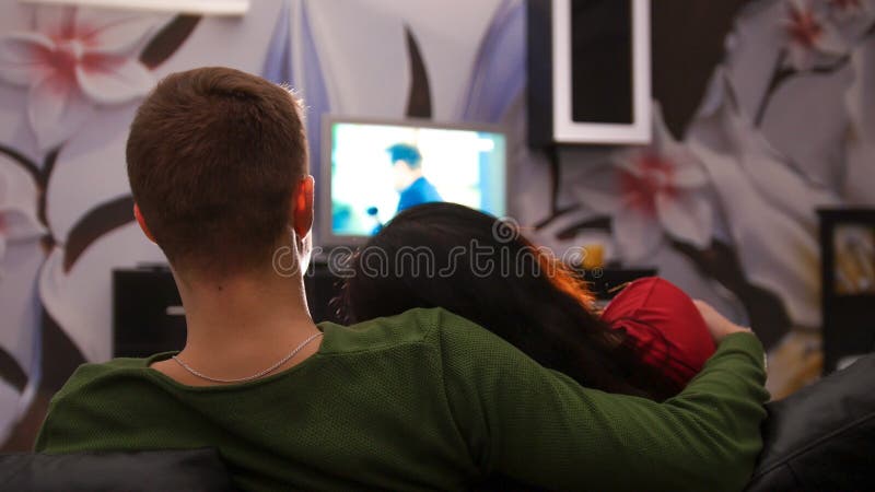 Jonge leuke paarzitting op een laag die op TV samen letten De vrouw legt op haar vriendschouder stock foto Jonge leuke paarzitting op een laag die op TV samen letten De vrouw legt op haar vriendschouder stock foto
