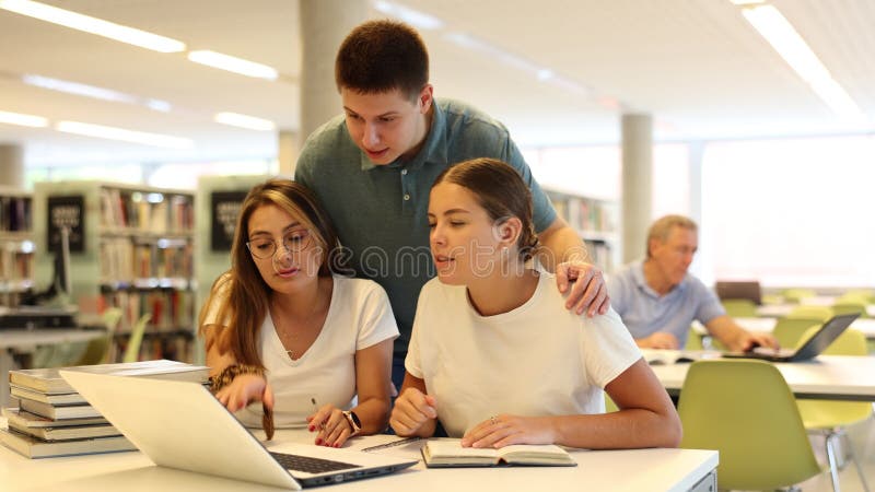 Jonge mannelijke bibliothecaris die advies geeft aan positieve vrouwelijke studenten die een laptop in de bibliotheek gebruiken
