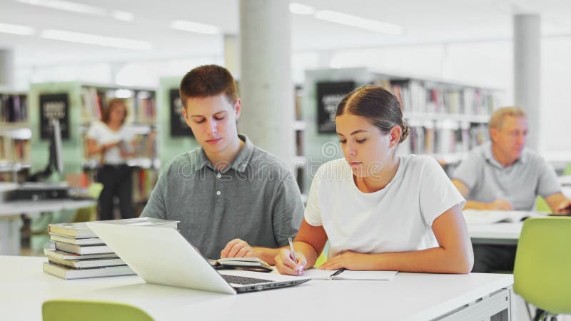 Jonge mannelijke bibliothecaris die informatie uitlegt voor onregelmatig vrouwtje terwijl ze een laptop in de bibliotheek gebruikt