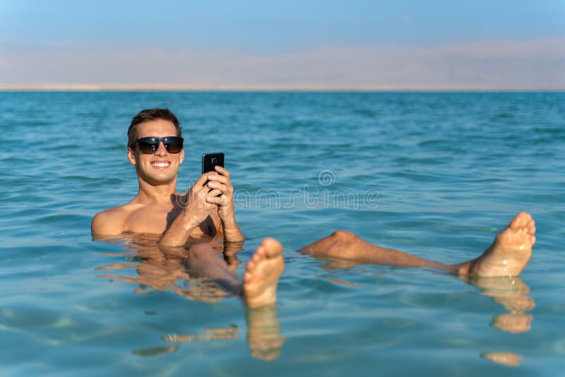 Jonge mens die op de waterspiegel van het dode overzees drijven en zijn smartphone gebruiken stock fotografie Jonge mens die op de waterspiegel van het dode overzees drijven en zijn smartphone gebruiken stock fotografie