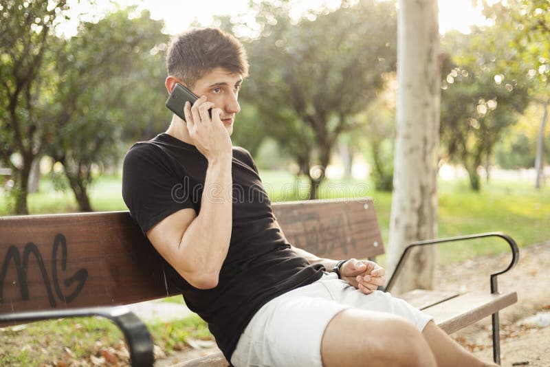 Jonge mens die telefoonzitting op een parkbank gebruiken stock afbeelding Jonge mens die telefoonzitting op een parkbank gebruiken stock afbeelding