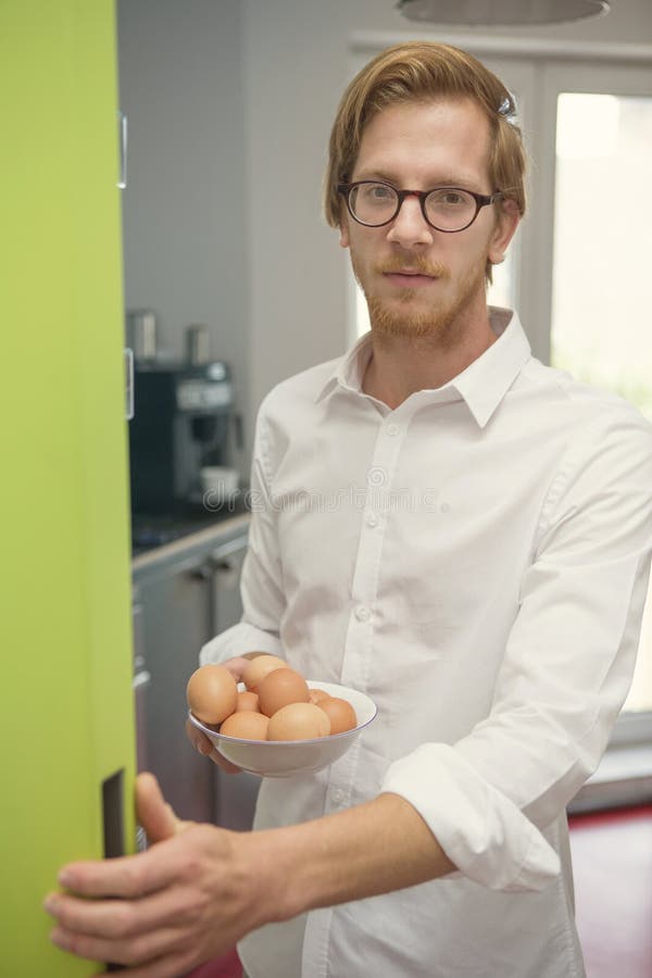 Jonge mens die zich bij ijskast met kom van eieren bevinden stock afbeelding Jonge mens die zich bij ijskast met kom van eieren bevinden stock afbeelding