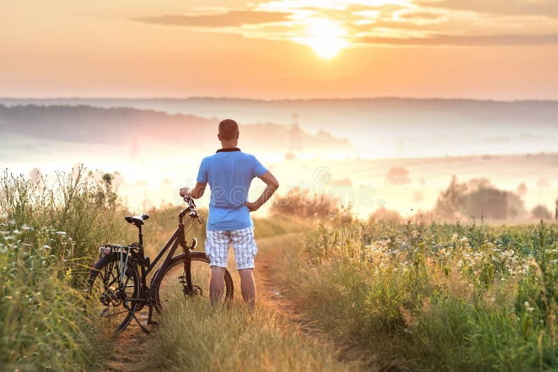 Jonge mens die zich dichtbij fiets in ochtendzonsopgang bevinden met wonderf stock afbeeldingen Jonge mens die zich dichtbij fiets in ochtendzonsopgang bevinden met wonderf stock afbeeldingen
