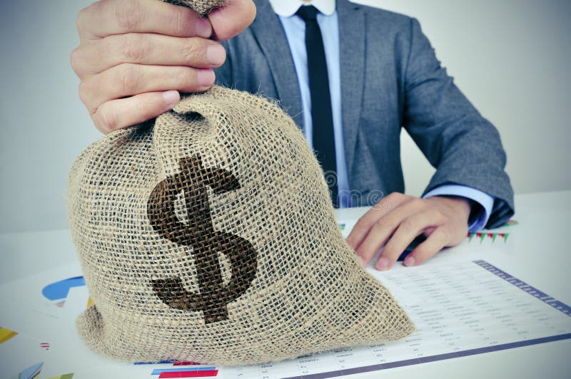 A young caucasian man wearing a gray suit seated at an office desk full of charts and financial balances holds a burlap money bag with the US dollar currency sign in his hand. A young caucasian man wearing a gray suit seated at an office desk full of charts and financial balances holds a burlap money bag with the US dollar currency sign in his hand