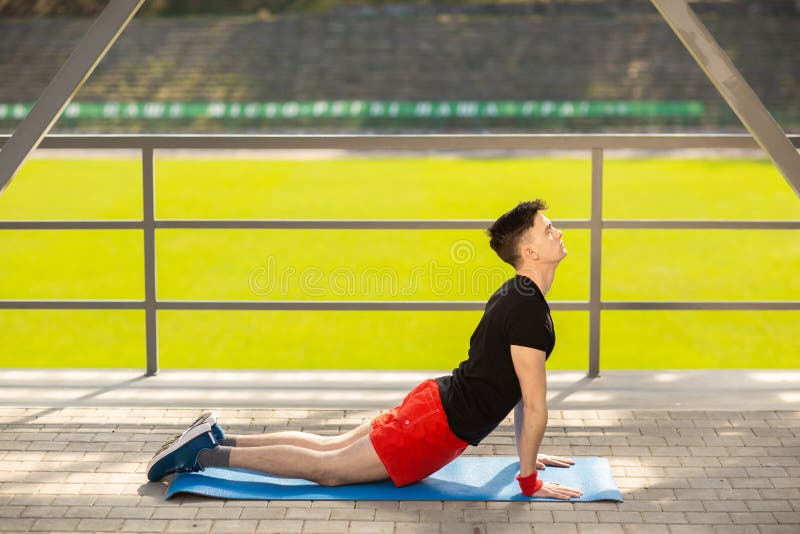 Jonge mens opleidingsyoga in openlucht De sportieve kerel maakt uitrekkende oefening op een blauwe yogamat, op de sportengrond stock foto Jonge mens opleidingsyoga in openlucht De sportieve kerel maakt uitrekkende oefening op een blauwe yogamat, op de sportengrond stock foto