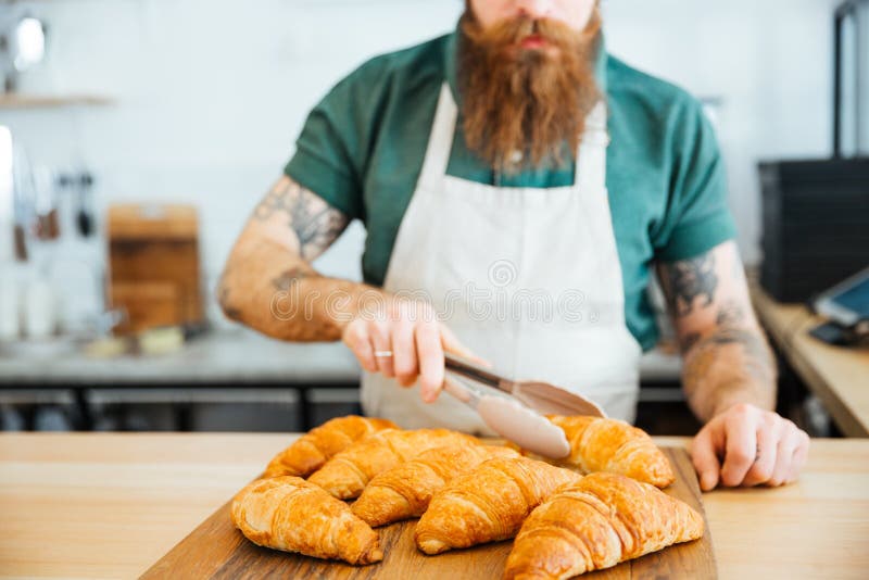 Jonge mensenbarista met baard die croissant nemen die tang met behulp van royalty-vrije stock afbeelding Jonge mensenbarista met baard die croissant nemen die tang met behulp van royalty-vrije stock afbeelding