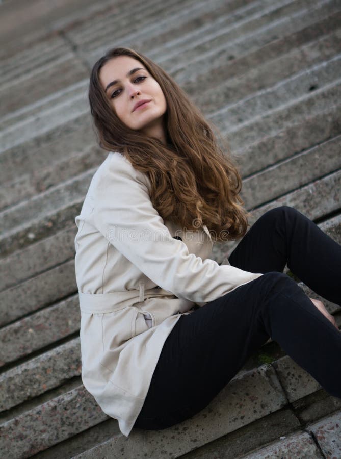 Jonge mooie vrouw die beige jasjezitting op concreet s dragen royalty-vrije stock foto's Jonge mooie vrouw die beige jasjezitting op concreet s dragen royalty-vrije stock foto's