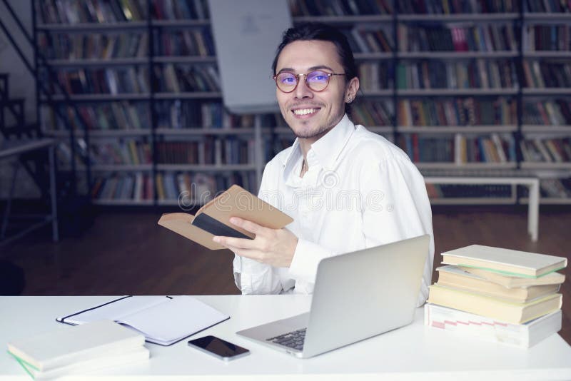 Jonge ondernemer of het universitaire student glimlachen, die aan laptop werken die een boek in een bibliotheek lezen