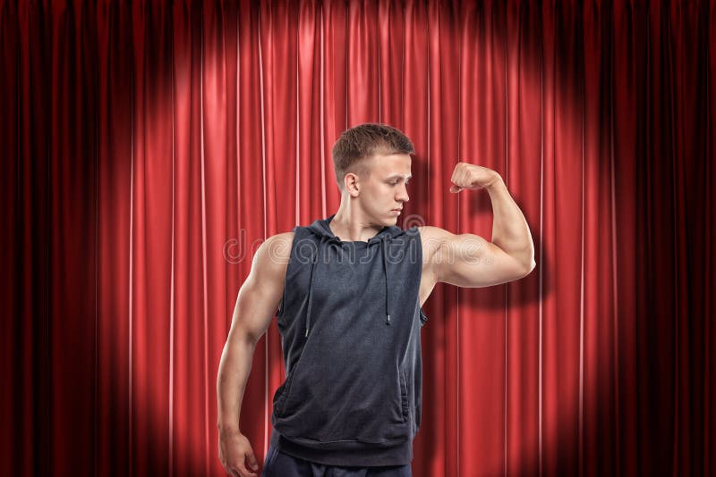Jonge spiermens in zwarte sportkleding die linkerwapenbicepsen op de rode achtergrond van stadiumgordijnen tonen royalty-vrije stock foto Jonge spiermens in zwarte sportkleding die linkerwapenbicepsen op de rode achtergrond van stadiumgordijnen tonen royalty-vrije stock foto