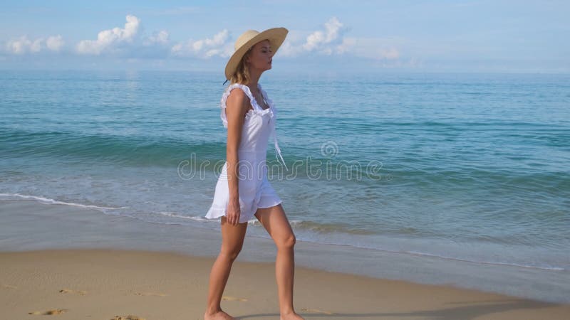 Jonge , vrij blonde , gelukkige , mooie vrouw in witte jurk en hoed die alleen loopt op een tropische zandstrand op de oceaan .