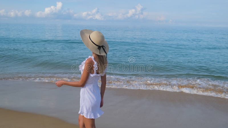 Jonge , vrij blonde , gelukkige , mooie vrouw in witte jurk en hoed die alleen loopt op een tropische zandstrand op de oceaan .
