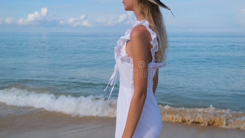 Jonge , vrij blonde , gelukkige , mooie vrouw in witte jurk en hoed die alleen loopt op een tropische zandstrand op de oceaan .