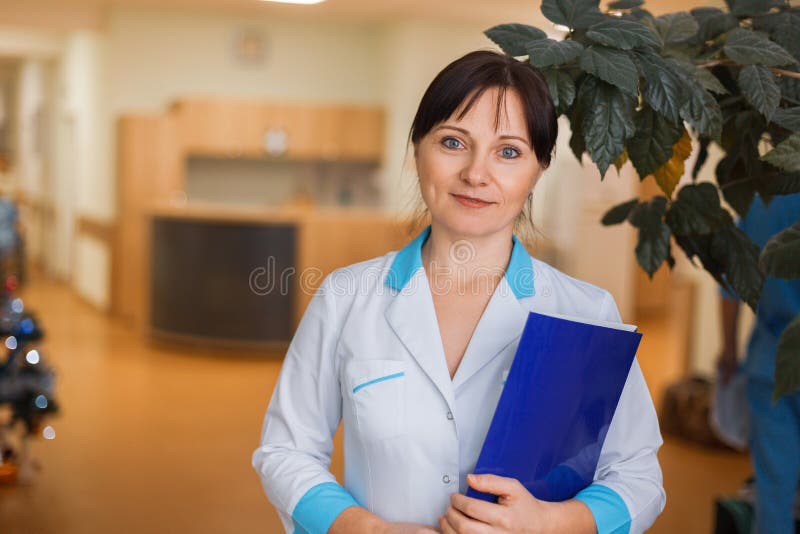 Jonge vrouw arts in medische witte kleren met een omslag voor documenten die zich in het ziekenhuis bevinden stock fotografie Jonge vrouw arts in medische witte kleren met een omslag voor documenten die zich in het ziekenhuis bevinden stock fotografie