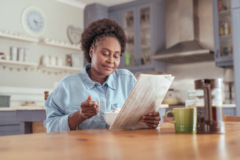 Jonge vrouw die de krant lezen terwijl thuis het eten van ontbijt stock afbeelding Jonge vrouw die de krant lezen terwijl thuis het eten van ontbijt stock afbeelding