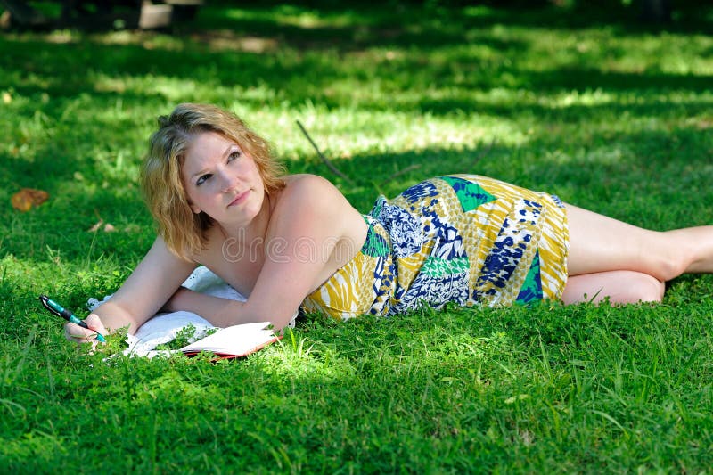 Attractive young woman in a brightly colored dress laying in the grass and writing in her journal or diary on a summery day in the park. Attractive young woman in a brightly colored dress laying in the grass and writing in her journal or diary on a summery day in the park