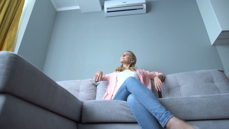 Jonge vrouw die op de bank zit onder de airconditioning, afkoelt op de hete zomer