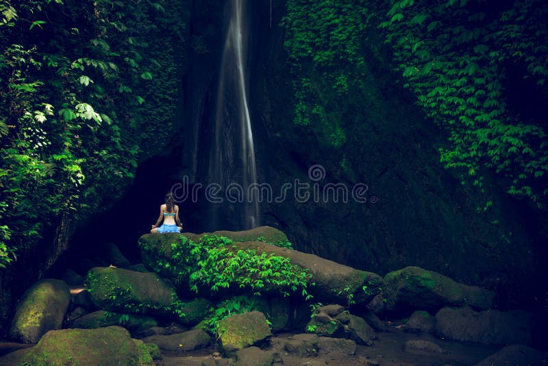 Jonge vrouw die op de rots zit en yoga bij waterval oefent Handen in gyan mudra Leke Leke waterval, Bali Achteraanzicht
