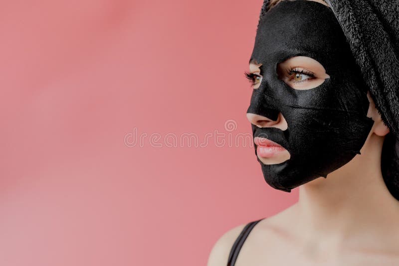 Young woman apply black cosmetic fabric facial mask on pink background. Face peeling mask with charcoal, spa beauty treatment, skincare, cosmetology. Close up. Young woman apply black cosmetic fabric facial mask on pink background. Face peeling mask with charcoal, spa beauty treatment, skincare, cosmetology. Close up.