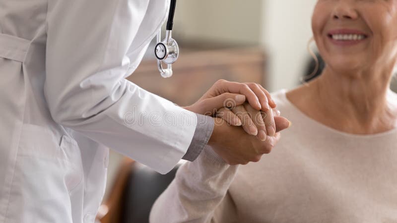 Doctor giving hope. Close up shot of young female physician leaning forward to smiling elderly lady patient holding her hand in palms. Woman caretaker in white coat supporting encouraging old person. Doctor giving hope. Close up shot of young female physician leaning forward to smiling elderly lady patient holding her hand in palms. Woman caretaker in white coat supporting encouraging old person