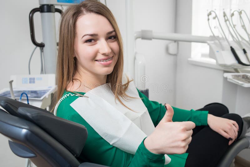 Jonge vrouwelijke patiënt met steunen op tanden die als tandvoorzitter zitten, en duimen na behandeling glimlachen tonen stock afbeeldingen Jonge vrouwelijke patiënt met steunen op tanden die als tandvoorzitter zitten, en duimen na behandeling glimlachen tonen stock afbeeldingen