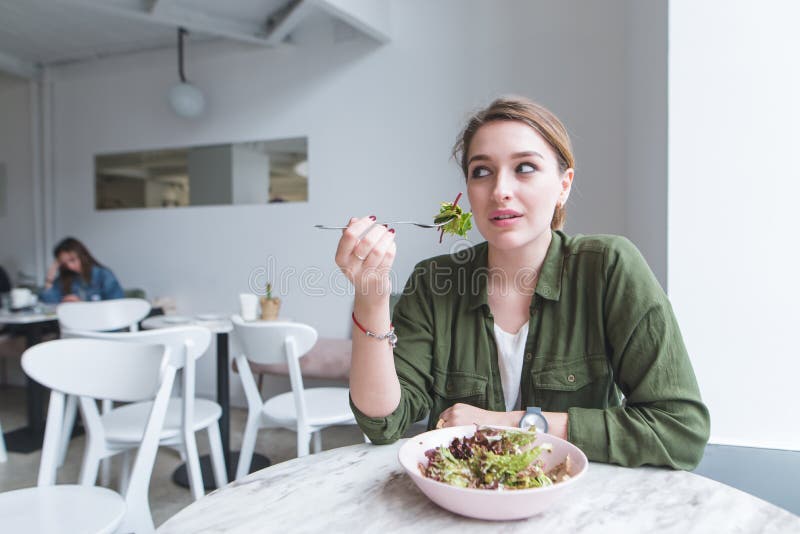 Jonge vrouwenzitting in een comfortabel licht restaurant met een vork in haar salade eten en handen die, die weg eruit zien stock fotografie Jonge vrouwenzitting in een comfortabel licht restaurant met een vork in haar salade eten en handen die, die weg eruit zien stock fotografie