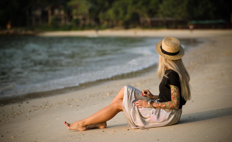 Jonge vrouwenzitting op de ochtend van het zandige strand Draagt een strohoed, een lange rok en een zwarte t-shirt De Gang van de royalty-vrije stock foto Jonge vrouwenzitting op de ochtend van het zandige strand Draagt een strohoed, een lange rok en een zwarte t-shirt De Gang van de royalty-vrije stock foto