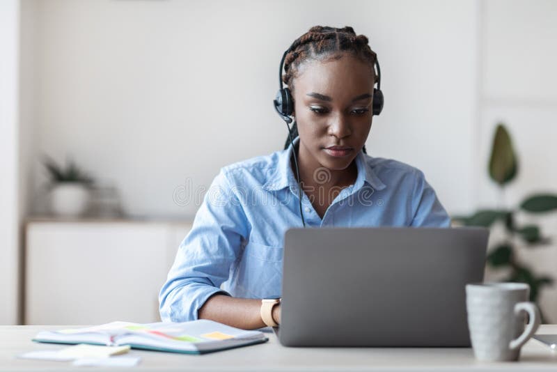 Jonge zwarte hotline operator vrouw in headset die werkt met laptop in kantoor