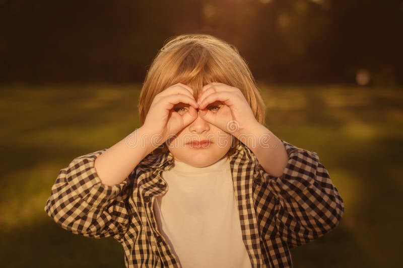 child boy in casual white t-shirt looking through fingers in binoculars gesture, observing distant with attentive look, watching afar. Hands make binoculars and eyes look through them. child boy in casual white t-shirt looking through fingers in binoculars gesture, observing distant with attentive look, watching afar. Hands make binoculars and eyes look through them