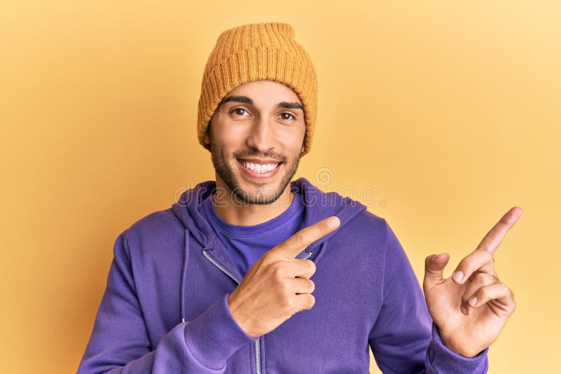 Young handsome man wearing wool winter hat smiling and looking at the camera pointing with two hands and fingers to the side. Young handsome man wearing wool winter hat smiling and looking at the camera pointing with two hands and fingers to the side