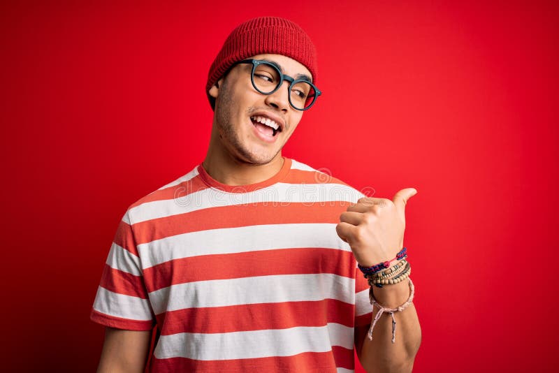 Young brazilian man wearing casual striped t-shirt and glasses over isolated red background smiling with happy face looking and pointing to the side with thumb up. Young brazilian man wearing casual striped t-shirt and glasses over isolated red background smiling with happy face looking and pointing to the side with thumb up