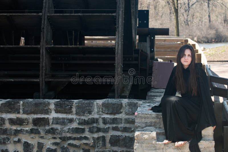 Young Woman with dark long hair in black robes in front of an Old Wooden Water Mill. Witches. Halloween concept. Witchcraft and Magic. Young Woman with dark long hair in black robes in front of an Old Wooden Water Mill. Witches. Halloween concept. Witchcraft and Magic.