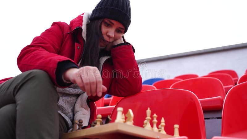 Jovem em roupas de inverno joga xadrez sentado nas arquibancadas do estádio  sozinho mulher de boné preto com xadrez no estádio esportivo em tempo  nublado