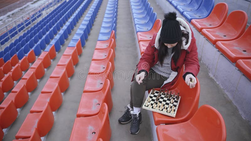 Jovem em roupas de inverno joga xadrez sentado nas arquibancadas do estádio  sozinho mulher de boné preto com xadrez no estádio esportivo em tempo  nublado