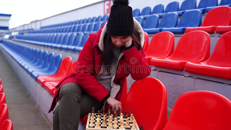 Jovem em roupas de inverno joga xadrez sentado nas arquibancadas do estádio  sozinho mulher de boné preto com xadrez no estádio esportivo em tempo  nublado