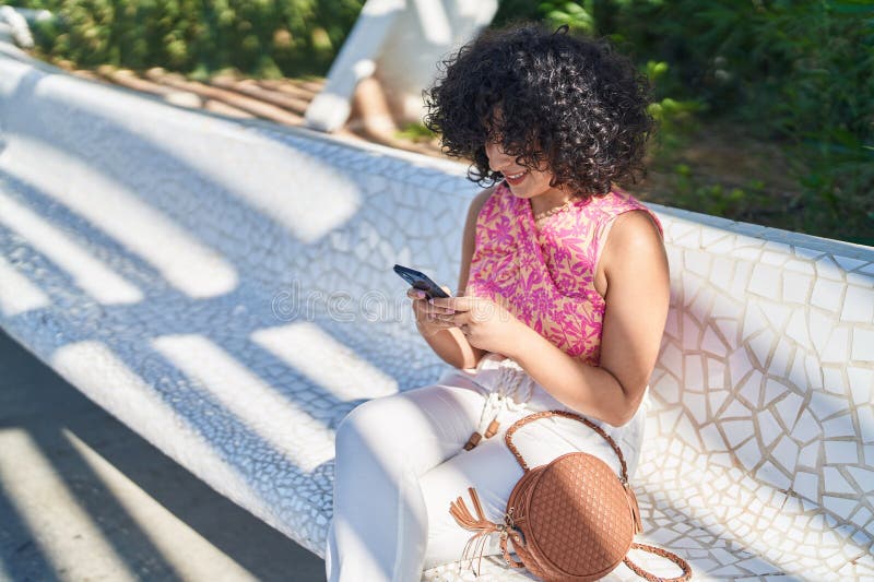 Jovem mulher do Médio Oriente a usar um smartphone sentada num banco num parque imagens de stock