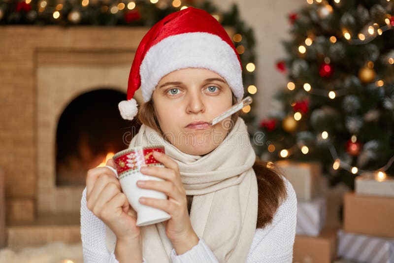 Young ill woman in santa hat, wrapped in scarf, measuring temperature and holding cup of warming drink, looks at camera with unhappy facial expression, posing indoor with fireplace and x-mas tree. Young ill woman in santa hat, wrapped in scarf, measuring temperature and holding cup of warming drink, looks at camera with unhappy facial expression, posing indoor with fireplace and x-mas tree