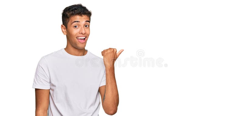 Young handsome african american man wearing casual white tshirt smiling with happy face looking and pointing to the side with thumb up. Young handsome african american man wearing casual white tshirt smiling with happy face looking and pointing to the side with thumb up