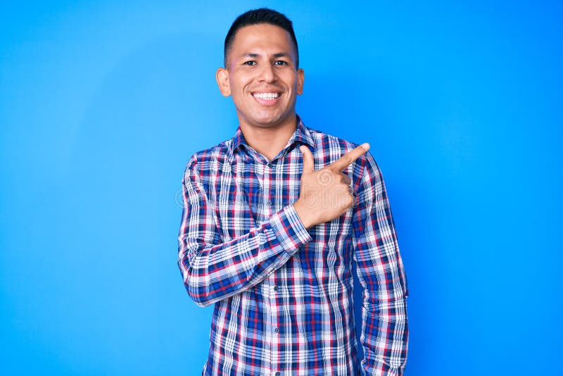 Young handsome latin man wearing casual clothes cheerful with a smile of face pointing with hand and finger up to the side with happy and natural expression on face. Young handsome latin man wearing casual clothes cheerful with a smile of face pointing with hand and finger up to the side with happy and natural expression on face