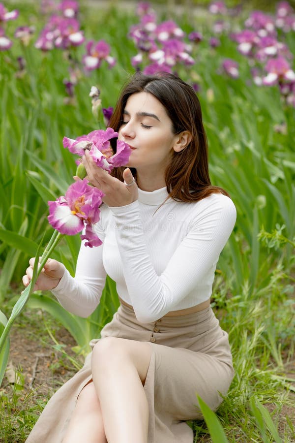 Side view of elegant, young female sitting on ground, holding flower, smelling with closed eyes. Concept of springtime. Side view of elegant, young female sitting on ground, holding flower, smelling with closed eyes. Concept of springtime.