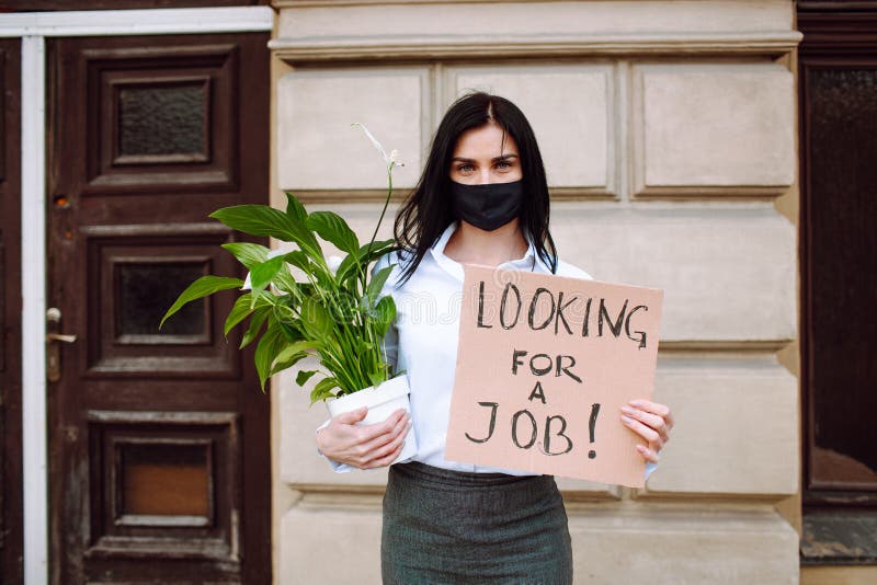 Jovem empresária segurando um cartaz em busca de emprego e escritório usando máscara preta. desemprego após o vírus corona