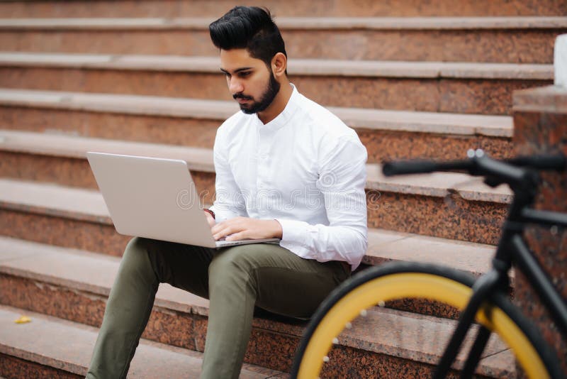 Jovem estudante indiano asiático a trabalhar no computador portátil enquanto se senta nas escadas da rua imagem de stock royalty free Jovem estudante indiano asiático a trabalhar no computador portátil enquanto se senta nas escadas da rua imagem de stock royalty free