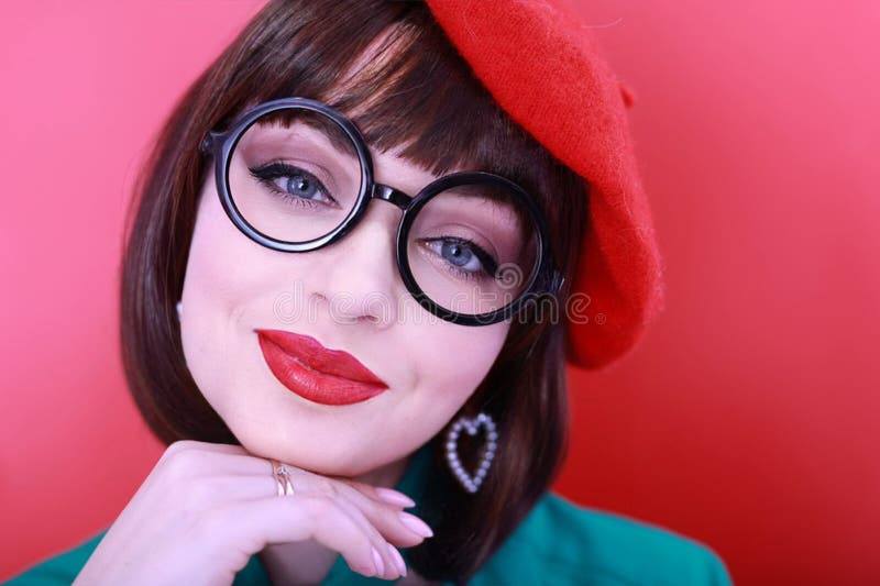 Young happy woman 30 years old wearing glasses, she wears a green grunt and a red hat, shows emotions, celebrates, clenching her fists, says yes, isolated on a plain red background, studio portrait. People lifestyle concept. Young happy woman 30 years old wearing glasses, she wears a green grunt and a red hat, shows emotions, celebrates, clenching her fists, says yes, isolated on a plain red background, studio portrait. People lifestyle concept