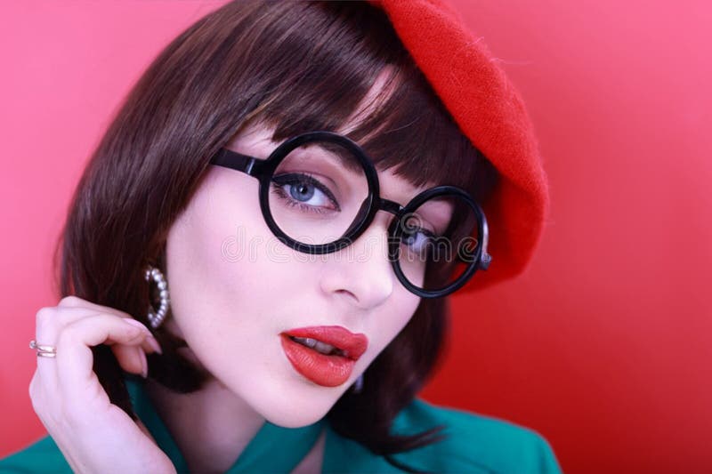 Young happy woman 30 years old wearing glasses, she wears a green grunt and a red hat, shows emotions, celebrates, clenching her fists, says yes, isolated on a plain red background, studio portrait. People lifestyle concept. Young happy woman 30 years old wearing glasses, she wears a green grunt and a red hat, shows emotions, celebrates, clenching her fists, says yes, isolated on a plain red background, studio portrait. People lifestyle concept