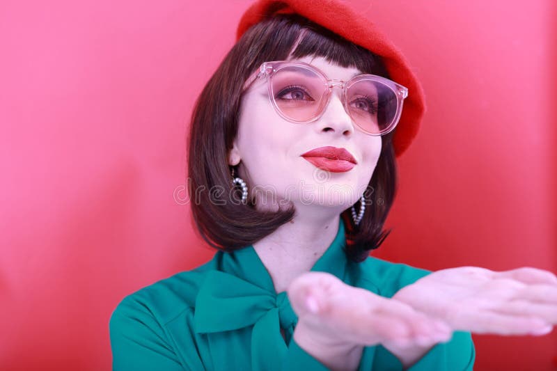 Young happy woman 30 years old wearing glasses, she wears a green grunt and a red hat, shows emotions, celebrates, clenching her fists, says yes, isolated on a plain red background, studio portrait. People lifestyle concept. Young happy woman 30 years old wearing glasses, she wears a green grunt and a red hat, shows emotions, celebrates, clenching her fists, says yes, isolated on a plain red background, studio portrait. People lifestyle concept