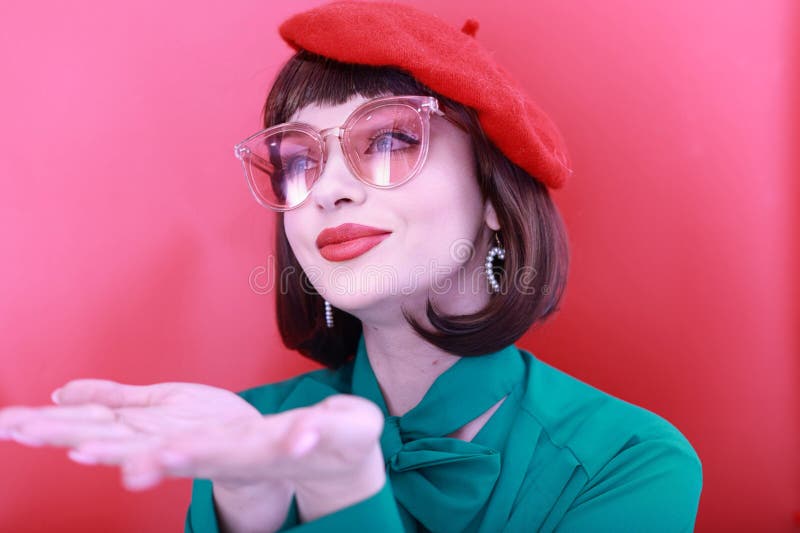 Young happy woman 30 years old wearing glasses, she wears a green grunt and a red hat, shows emotions, celebrates, clenching her fists, says yes, isolated on a plain red background, studio portrait. People lifestyle concept. Young happy woman 30 years old wearing glasses, she wears a green grunt and a red hat, shows emotions, celebrates, clenching her fists, says yes, isolated on a plain red background, studio portrait. People lifestyle concept