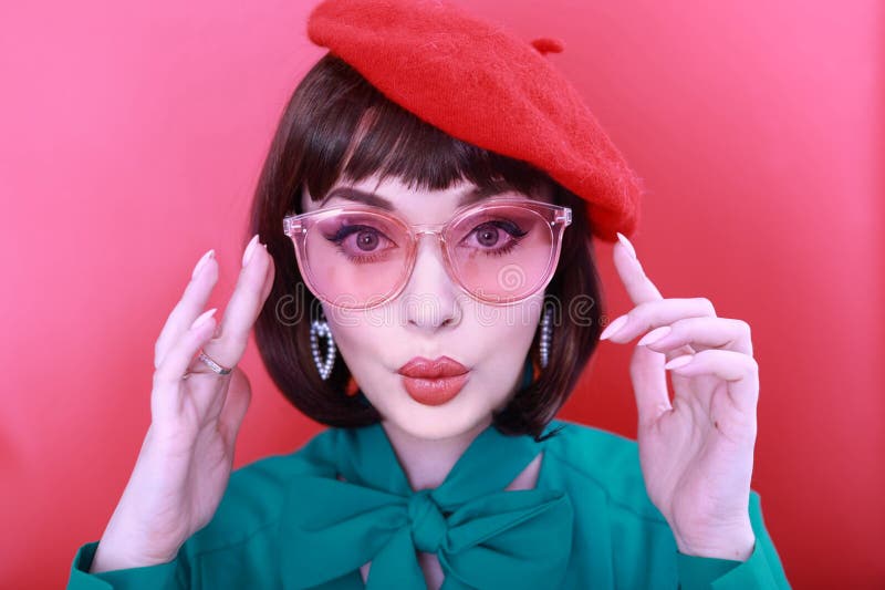 Young happy woman 30 years old wearing glasses, she wears a green grunt and a red hat, shows emotions, celebrates, clenching her fists, says yes, isolated on a plain red background, studio portrait. People lifestyle concept. Young happy woman 30 years old wearing glasses, she wears a green grunt and a red hat, shows emotions, celebrates, clenching her fists, says yes, isolated on a plain red background, studio portrait. People lifestyle concept