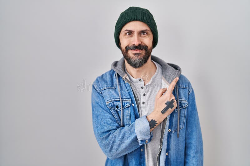 Young hispanic man with tattoos wearing wool cap cheerful with a smile of face pointing with hand and finger up to the side with happy and natural expression on face. Young hispanic man with tattoos wearing wool cap cheerful with a smile of face pointing with hand and finger up to the side with happy and natural expression on face