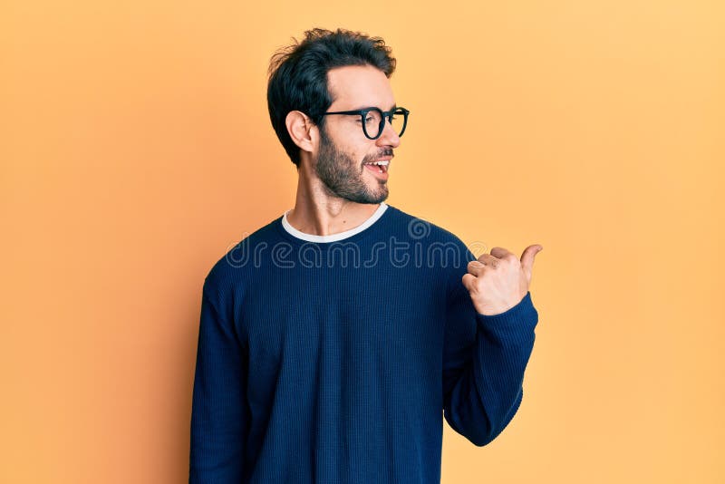 Young hispanic man wearing casual clothes and glasses smiling with happy face looking and pointing to the side with thumb up. Young hispanic man wearing casual clothes and glasses smiling with happy face looking and pointing to the side with thumb up