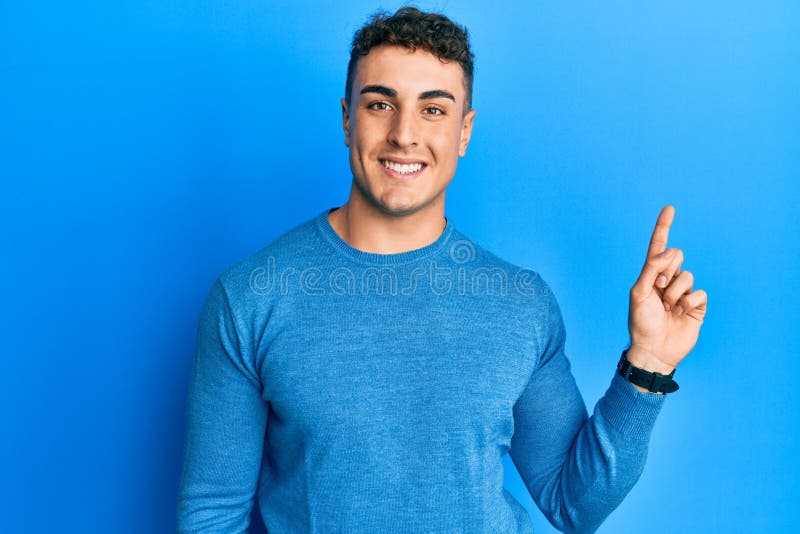 Hispanic young man wearing casual winter sweater with a big smile on face, pointing with hand finger to the side looking at the camera. Hispanic young man wearing casual winter sweater with a big smile on face, pointing with hand finger to the side looking at the camera