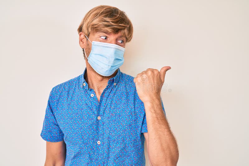 Young blond man wearing medical mask pointing thumb up to the side smiling happy with open mouth. Young blond man wearing medical mask pointing thumb up to the side smiling happy with open mouth